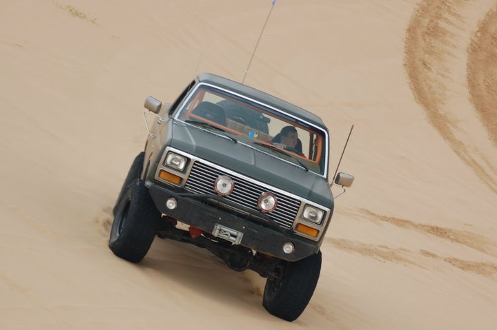 Dark green Ford Bronco with aftermarket bumper and winch driving up a sand dune. The vehicle has large tires and a CB antenna