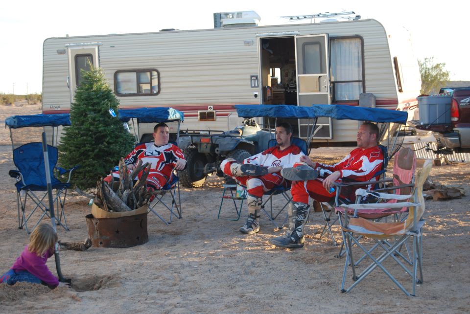 Three men in motocross gear sit in chairs around a fire pit near a camper and an ATV