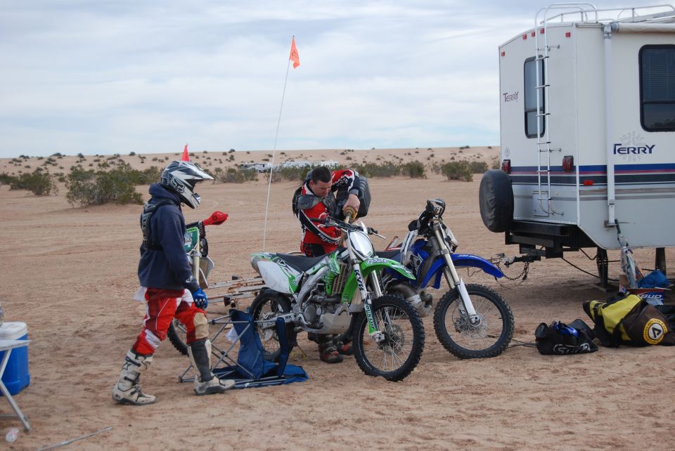 Two dirt bikes and a white RV parked on sand dunes under a cloudy sky