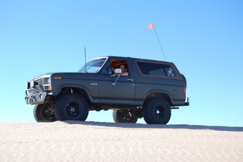 1982 Ford Bronco - A dark gray 1980 Ford Bronco with large off-road tires navigates sandy terrain under a clear blue sky.