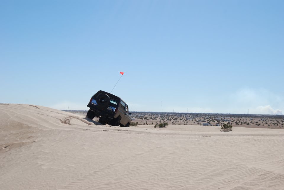 Bronco climbing sand dune with flag on antenna. Desert landscape with wind turbines in background