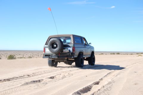 1982 Ford Bronco - A gray Ford Bronco kicks up sand as it drives through a desert landscape.