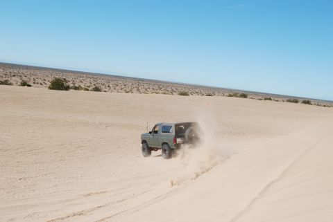 1982 Ford Bronco - A 1982 Ford Bronco navigates a sandy terrain, its tires kicking up a cloud of dust against a clear blue sky.