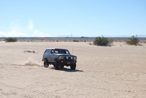 1982 Ford Bronco - A gray SUV navigates a sandy terrain under a clear blue sky.