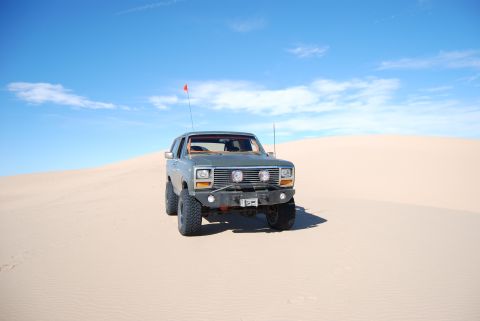 1982 Ford Bronco - This 1980s Ford Bronco, modified for off-road use, navigates a sandy desert landscape under a clear blue sky.