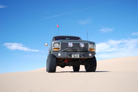 1982 Ford Bronco - This modified truck sits on a sandy incline, ready for off-road adventure under a bright blue sky with scattered clouds.