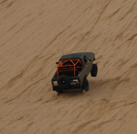 1982 Ford Bronco - A pickup truck with an orange roll cage navigates a sandy incline, kicking up dust.
