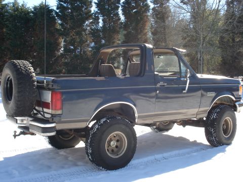 1988 Ford Bronco - Rear three-quarter view of a lifted dark blue Ford Bronco with swing-away spare tire, parked on snow with trees behind.