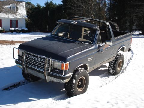 1988 Ford Bronco - A dark blue 1980s Ford Bronco with its top removed, sitting on snow-covered ground in front of a white house with red-trimmed windows. The…
