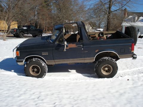 1988 Ford Bronco - Driver-side profile of a lifted dark blue Ford Bronco with the hardtop removed, showing the exposed roll bar, tan interior and bare rear tu…