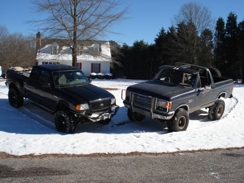 1988 Ford Bronco - Two lifted black Fords parked side by side on a snowy driveway in front of a white farmhouse: a black extended-cab Ranger with a front winc…