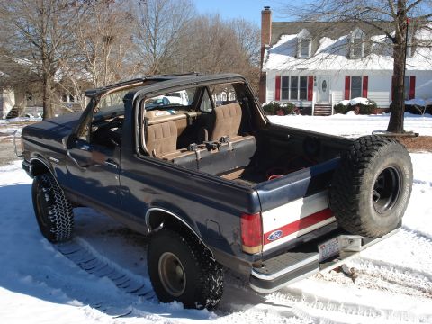 1988 Ford Bronco - Rear three-quarter view of a dark blue 1987-91-style full-size Ford Bronco with the top removed exposing tan seats, a rear-mounted Mud Terr…