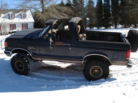 1988 Ford Bronco - Side view of a dark blue lifted Ford Bronco with the rear top removed, parked in snow by a house.