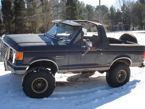 1988 Ford Bronco - A dark blue 1988 Ford Bronco with the removable hardtop off, shown in side/3q-front view on snowy ground. It rides on a suspension lift wit…