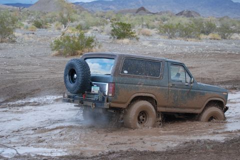 1982 Ford Bronco - A green Ford Bronco navigates through a muddy terrain, with its rear tire mounted externally.