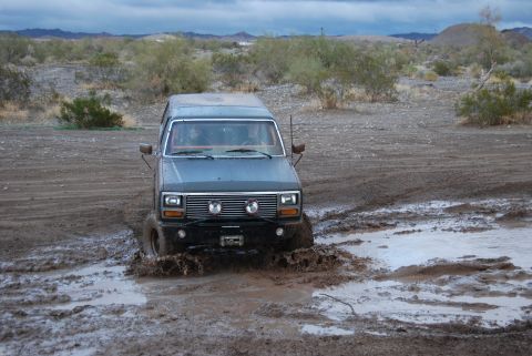 1982 Ford Bronco - A gray Ford Bronco navigates a muddy terrain, kicking up water and mud.