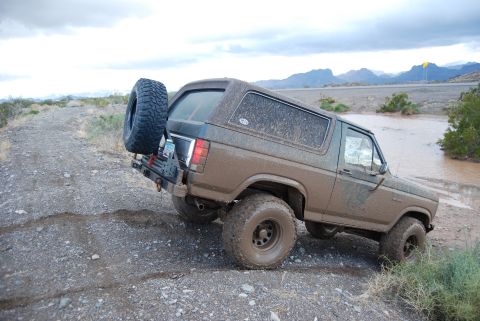 1982 Ford Bronco - Muddy 1982 Ford Bronco navigates a water crossing on a dirt road with mountains in the background.