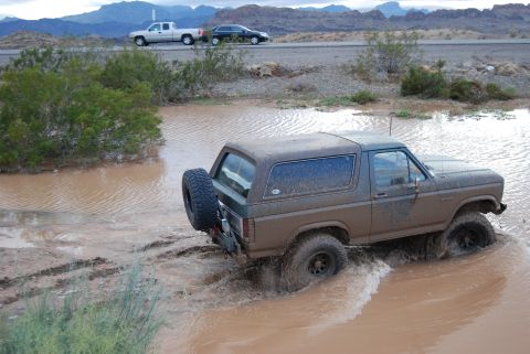 1982 Ford Bronco - A Ford Bronco navigates a muddy off-road terrain, with other vehicles visible on a distant road.