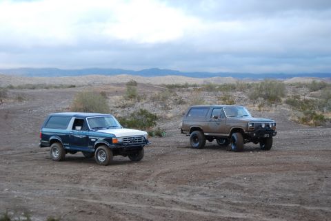1982 Ford Bronco - Two Ford Broncos, one blue and white and the other brown, are parked in a dusty desert landscape under a cloudy sky.