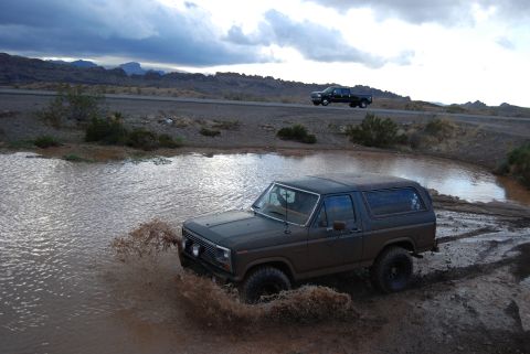 1982 Ford Bronco - Off-road Bronco navigates a muddy terrain under a cloudy sky, with another truck visible on the distant road.