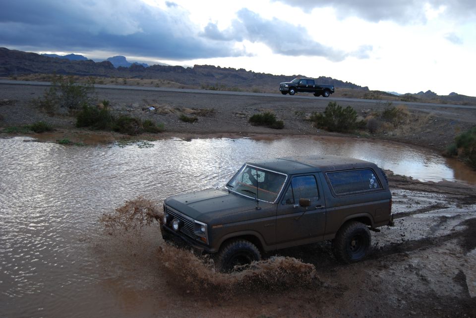 Dark 1980s Ford Bronco splashing through a muddy puddle off-road, with a black pickup truck driving on a road in the background