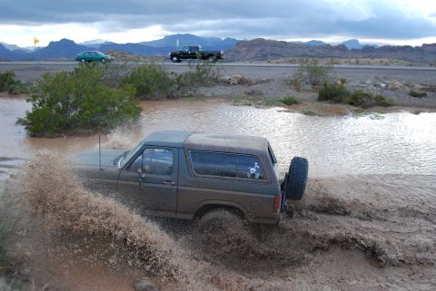 1982 Ford Bronco - This Ford Bronco is navigating a muddy off-road area, kicking up a lot of water and dirt.