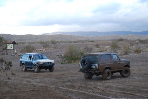 1982 Ford Bronco - Two Ford Broncos are parked in a dusty, desert landscape under a cloudy sky.