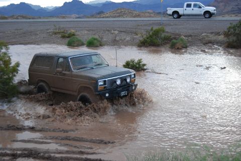 1982 Ford Bronco - A brown 1982 Ford Bronco navigates a muddy water obstacle, with a white pickup truck visible on the road in the background.