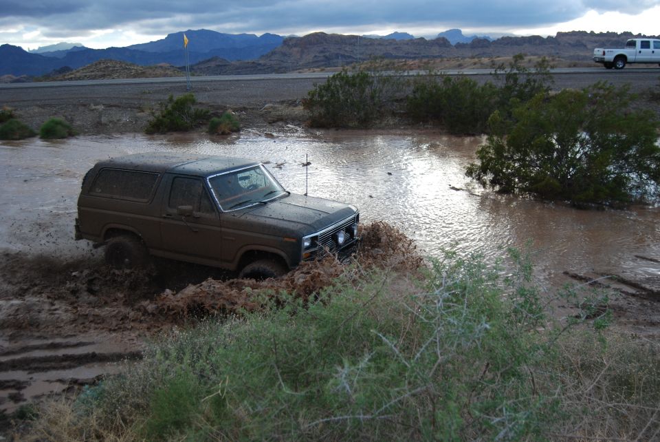 Brown Ford Bronco driving through muddy water with mountains in background
