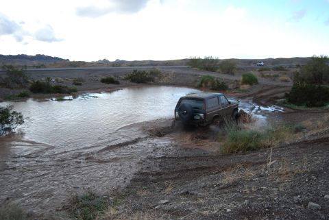 1982 Ford Bronco - A brown Ford Bronco navigates a muddy puddle in a dry, arid landscape.