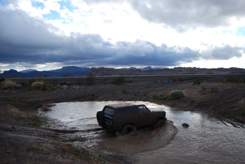 1982 Ford Bronco - A dark brown SUV splashes through a muddy puddle in a , outdoor landscape under a cloudy sky.
