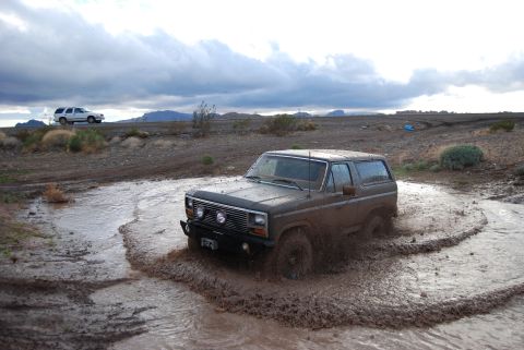 1982 Ford Bronco - A brown Ford Bronco navigates a muddy terrain, with a second vehicle in the distance under a cloudy sky.