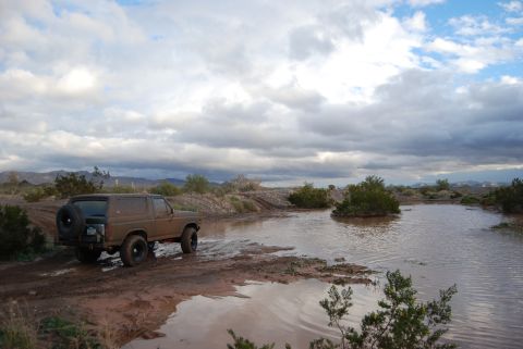 1982 Ford Bronco - A brown 1982 Ford Bronco navigates a muddy, water-filled terrain under a cloudy sky.