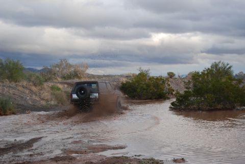 1982 Ford Bronco - Vehicle splashes through muddy water under a cloudy sky.