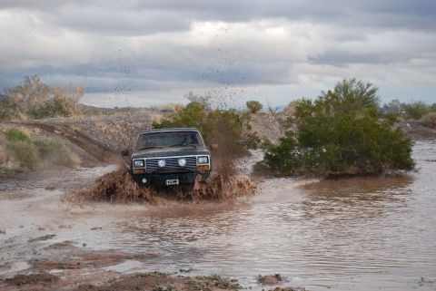 1982 Ford Bronco - A Jeep Cherokee XJ navigates a muddy off-road trail, kicking up a large spray.