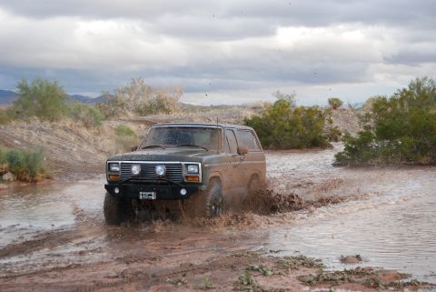 1982 Ford Bronco - A dark SUV navigates a muddy off-road path, kicking up water and dirt.