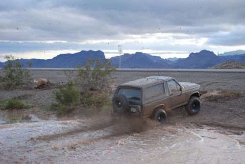 1982 Ford Bronco - A 1980s Ford Bronco kicks up mud and water while navigating an off-road trail under a cloudy sky.