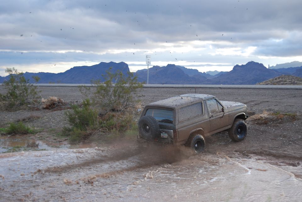 Muddy Ford Bronco driving through a water-filled dirt track with mountains in the background