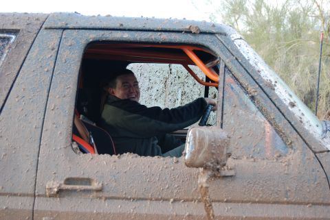1982 Ford Bronco - A man grins from the driver's seat of a heavily mud-splattered vehicle, its interior an orange roll cage.