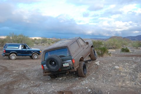 1982 Ford Bronco - Two Ford Broncos navigate a rocky, off-road trail under a cloudy sky.