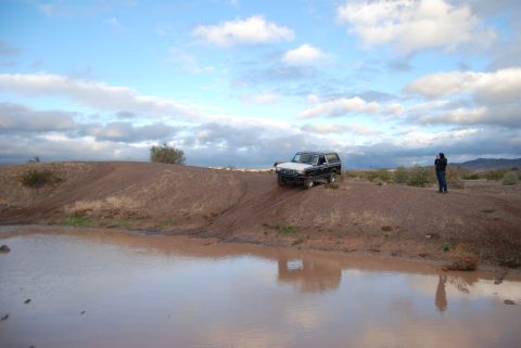 1982 Ford Bronco - A 1982 Ford Bronco navigates a dirt embankment near a body of water under a cloudy sky.