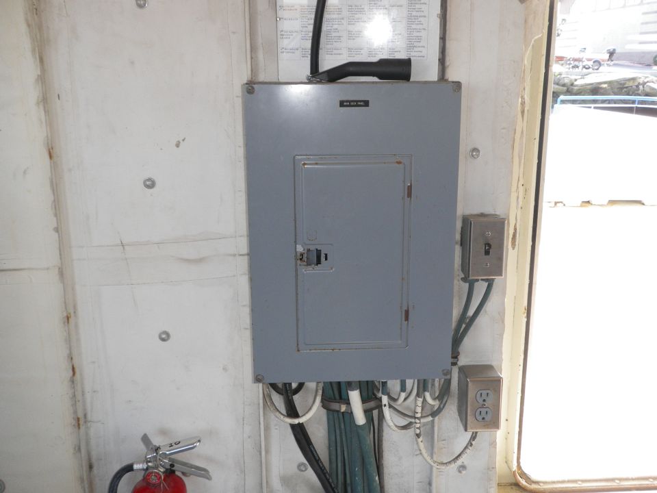 Gray electrical panel with a closed door, mounted on a concrete wall next to a fire extinguisher