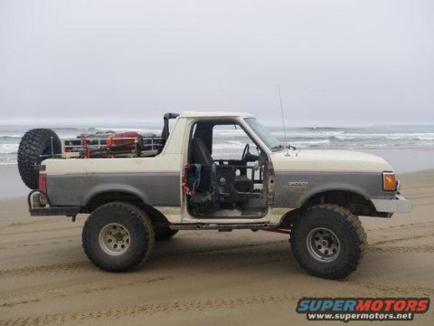 1991 Ford Bronco - A two-tone Ford Bronco sits on a beach with its doors off and a spare tire mounted at the rear.