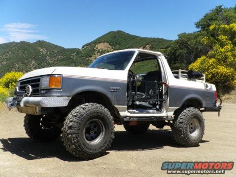 1991 Ford Bronco - This lifted 1991 Ford Bronco features large off-road tires and an open driver's side door, set against a backdrop of rolling hills.