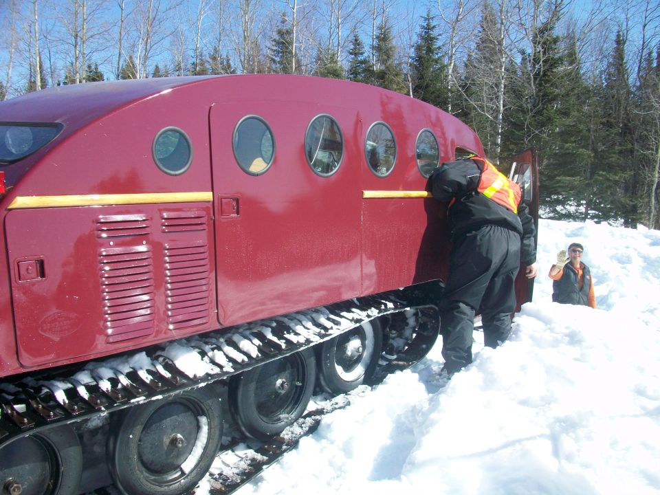 Red tracked snow vehicle with round windows in snow, person in orange vest at open door