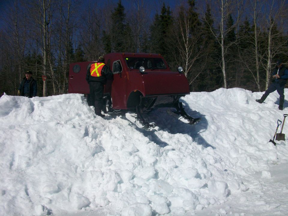 Maroon enclosed tracked snow vehicle on a snowbank with people nearby