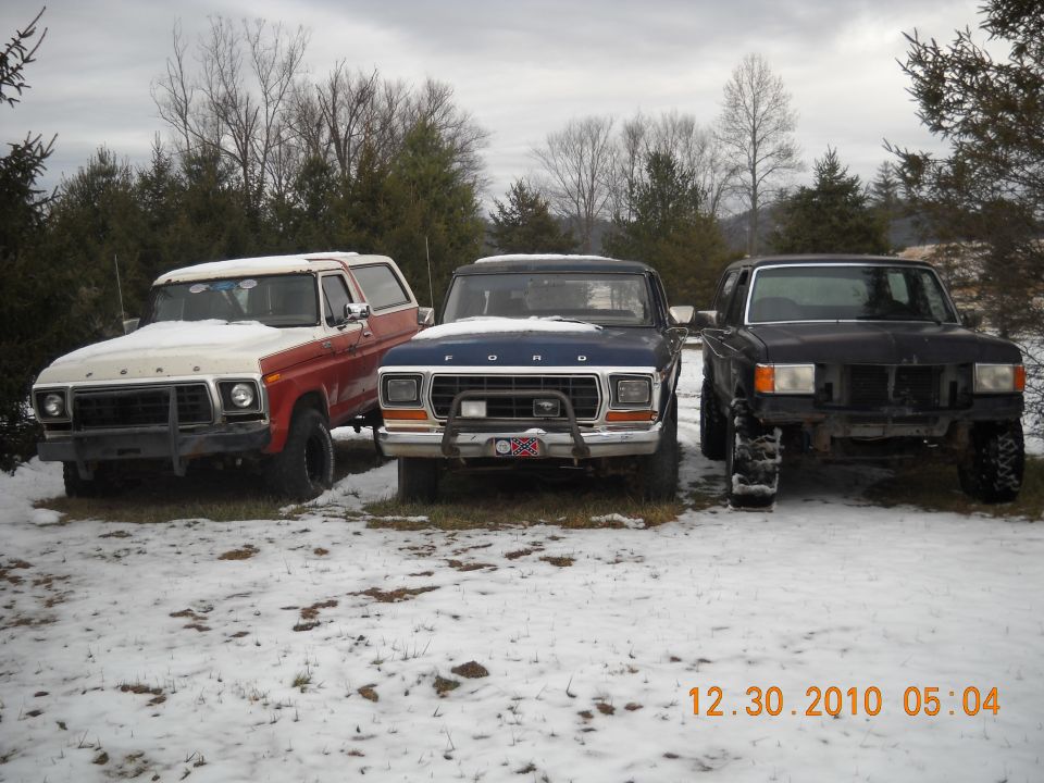 Three Ford Broncos parked in a snowy field. The one on the left is red and white, the middle one is blue, and the one on the right is black