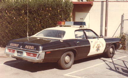 1991 Ford LTD Crown Victoria - Black-and-white 1970s Dodge Coronet highway patrol sedan seen from the rear three-quarter in a parking lot beside a hedge, with "HIGHWAY PA…