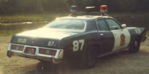 1991 Ford LTD Crown Victoria - Weathered dark blue and white 1970s Plymouth police sedan viewed from the rear three-quarter on a dirt lot, numbered 87 on the quarter pane…