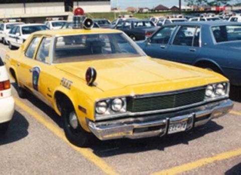 1991 Ford LTD Crown Victoria - A yellow mid-1970s Plymouth full-size police sedan sits in a parking lot, front three-quarter view, with a single red roof beacon, siren sp…